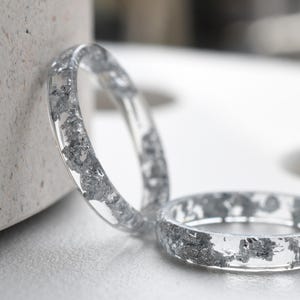 Puede incluir: Dos anillos de resina transparente con destellos plateados incrustados en el interior. Los anillos están apilados uno encima del otro.
