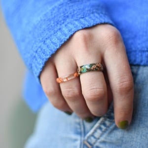 May include: Two rings are displayed. One ring is clear with orange accents. The other ring is green with copper-colored accents. The rings are on a finger, with a blue sweater and denim jeans in the background.