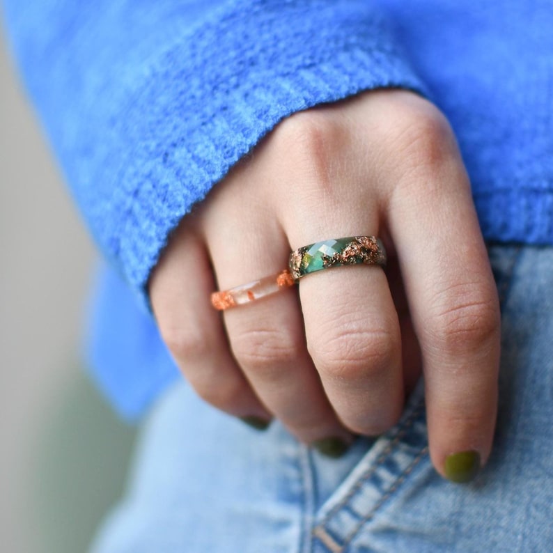 May include: Two rings are displayed. One ring is clear with orange accents. The other ring is green with copper-colored accents. The rings are on a finger, with a blue sweater and denim jeans in the background.