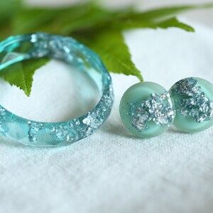 Puede incluir: Un conjunto de joyas de resina turquesa con escamas plateadas. El conjunto incluye un anillo y un par de pendientes.