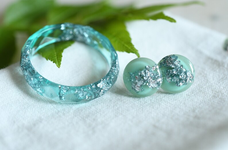 Puede incluir: Un conjunto de joyas de resina turquesa con escamas plateadas. El conjunto incluye un anillo y un par de pendientes.