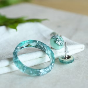 Puede incluir: Un conjunto de joyas de resina turquesa con detalles de purpurina plateada. El conjunto incluye una pulsera de brazalete y un par de pendientes de bot&oacute;n.