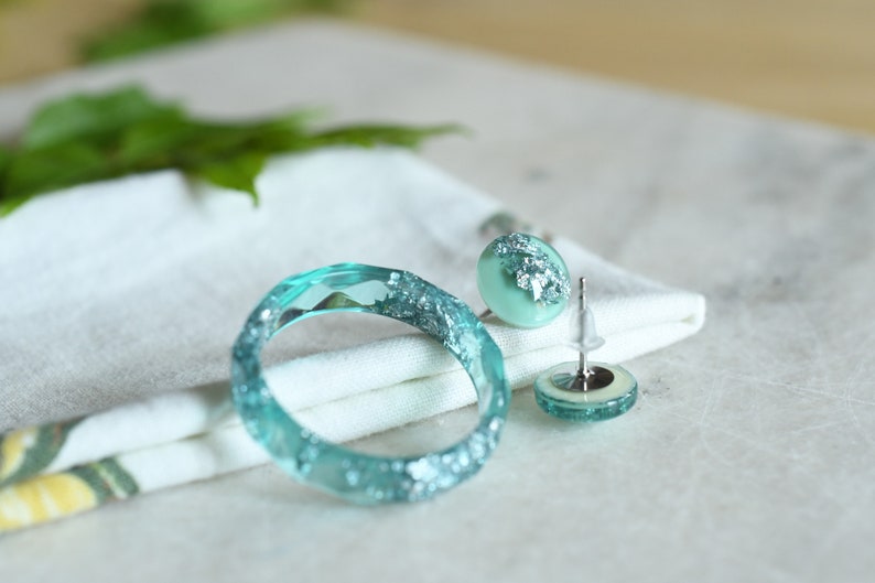 Puede incluir: Un conjunto de joyas de resina turquesa con detalles de purpurina plateada. El conjunto incluye una pulsera de brazalete y un par de pendientes de bot&oacute;n.
