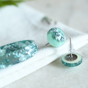 Puede incluir: Un conjunto de joyas de resina azul turquesa con escamas plateadas. El conjunto incluye un anillo y un par de pendientes.