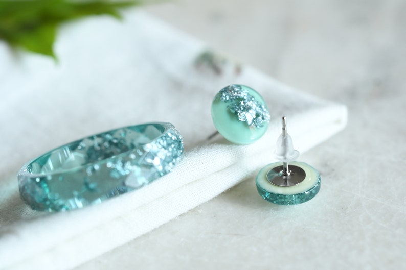 Puede incluir: Un conjunto de joyas de resina azul turquesa con escamas plateadas. El conjunto incluye un anillo y un par de pendientes.