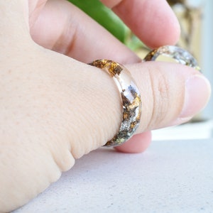 K&ouml;nnte beinhalten: Zwei transparente Resinringe mit eingebetteten goldenen und silbernen Flocken. Die Ringe werden an einem Finger getragen.