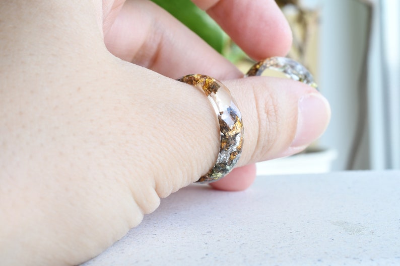 K&ouml;nnte beinhalten: Zwei transparente Resinringe mit eingebetteten goldenen und silbernen Flocken. Die Ringe werden an einem Finger getragen.
