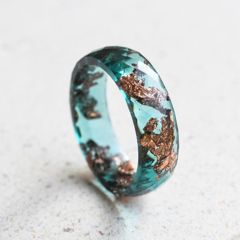 May include: A faceted teal resin ring with copper leaf inclusions. The ring has a clear, transparent appearance, with the copper leaf adding a metallic contrast. The ring is a unique piece of jewelry.