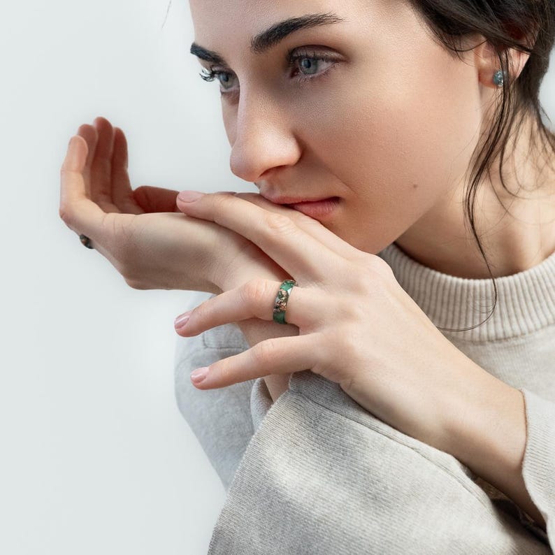 May include: A close-up of a ring with a green stone and silver accents. The ring is on a person's finger. The person is wearing a light gray sweater.