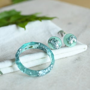 Puede incluir: Un conjunto de joyas de color azul turquesa con detalles de purpurina plateada. El conjunto incluye una pulsera de brazalete y un par de pendientes de bot&oacute;n.