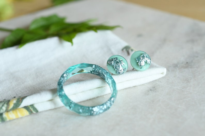 Puede incluir: Un conjunto de joyas de color azul turquesa con detalles de purpurina plateada. El conjunto incluye una pulsera de brazalete y un par de pendientes de bot&oacute;n.