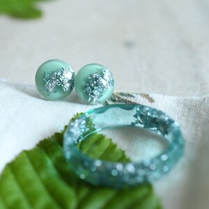 Puede incluir: Un conjunto de pendientes de resina verde turquesa y un anillo a juego con detalles de hojas de plata. Las joyas se muestran sobre una superficie de tela blanca con hojas verdes.