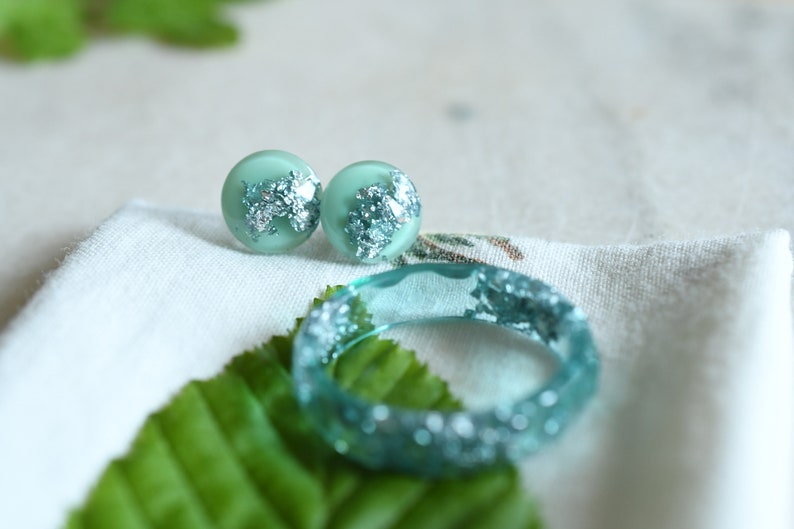 Puede incluir: Un conjunto de pendientes de resina verde turquesa y un anillo a juego con detalles de hojas de plata. Las joyas se muestran sobre una superficie de tela blanca con hojas verdes.