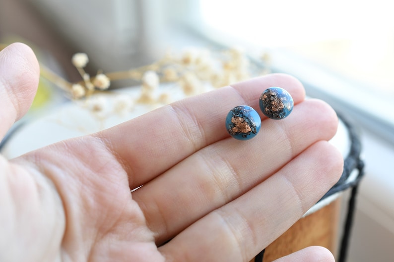 Puede incluir: Un par de pendientes de bot&oacute;n redondos con un dise&ntilde;o azul y cobre. Los pendientes est&aacute;n hechos de resina y tienen un acabado met&aacute;lico.