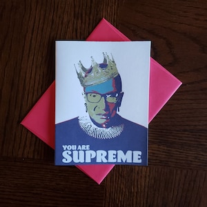 May include: A greeting card with a portrait of Ruth Bader Ginsburg wearing a crown. The text "You are Supreme" is printed in bold, colorful letters below the portrait.