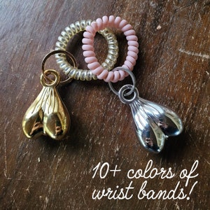Può includere: Due paia di braccialetti colorati, un paio è dorato e l'altro è argentato. I braccialetti sono attaccati a portachiavi con un ciondolo su ogni portachiavi. Il testo "10+ colori di braccialetti!" è scritto sotto l'immagine.