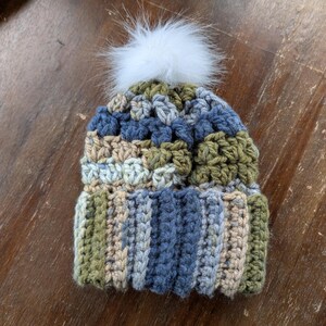 May include: A crocheted beanie hat with a white pom pom. The hat is made with a variety of colors, including green, blue, and beige.