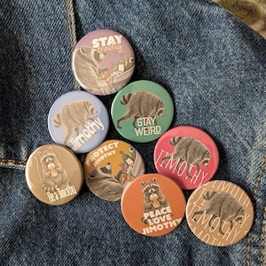 May include: A collection of eight circular pin-back buttons featuring illustrations of raccoons. The buttons display various colors including purple, blue, green, pink, orange, and tan, with text such as Stay Trashy, Jimothy, Stay Weird, Be a Jimothy, Protect Jimothy, and Peace Love Jimothy.