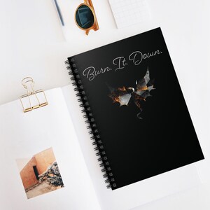 以下が含まれることがあります： 「Burn. It. Down.」の白い筆記体で書かれた黒いリングノート。炎のアクセントが付いた様式化されたドラゴンのグラフィックがテキストの下に配置されています。ノートは白い表面に開かれています。