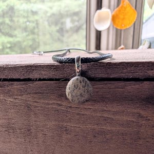 May include: A natural stone pendant necklace. The pendant is a rounded, textured stone in shades of gray and brown, suspended from a silver-colored bail. The necklace features a dark, braided leather cord. The background includes a wooden surface and a window.