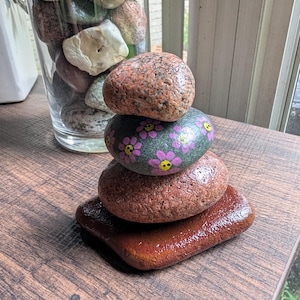 May include: A stack of four smooth, polished stones arranged vertically. The top stone is speckled pink and gray, the second is dark gray with painted pink flowers and yellow centers, the third is reddish-brown, and the base is a flat, rectangular reddish-brown stone.