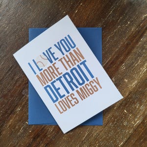May include: A white greeting card with the text "I LOVE YOU MORE THAN DETROIT LOVES MIGGY" in blue and brown lettering, with a baseball graphic. The card rests on a blue envelope, set against a wooden surface.