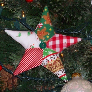 May include: A patchwork star ornament with red and white gingham, red and white stripes, green and white floral print, brown and green gingerbread print, and a green and white "Santa's Chocolate Shop" print. The star is hanging on a green Christmas tree.