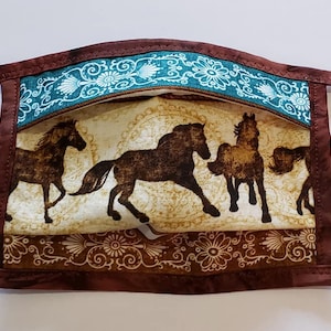 May include: A brown fabric face mask with a teal and white floral pattern. The mask features a design of brown horses running on a beige background.