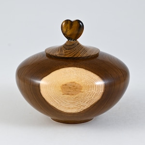 Puede incluir: Un tarro de madera hecho a mano con tapa y asa en forma de corazón. El tarro es de color marrón oscuro con un patrón de vetas de madera más claro en la tapa. El centro del tarro tiene un patrón de vetas de madera más claro. El asa es marrón.