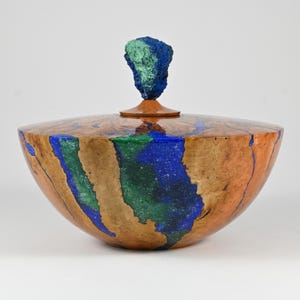 Peut inclure: Un bol en bois poli avec un couvercle surmonté d'un bouton texturé et brut. Le bol présente un grain de bois naturel brun clair entremêlé de sections incrustées aux teintes vibrantes de bleu profond et de vert minéral éclatant.