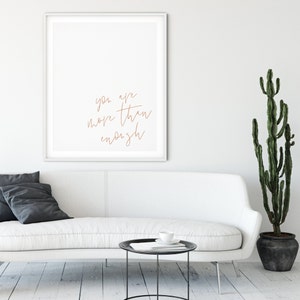 May include: A white couch with a black side table and a potted cactus plant. A framed print with the text "you are more than enough" hangs on the wall behind the couch.