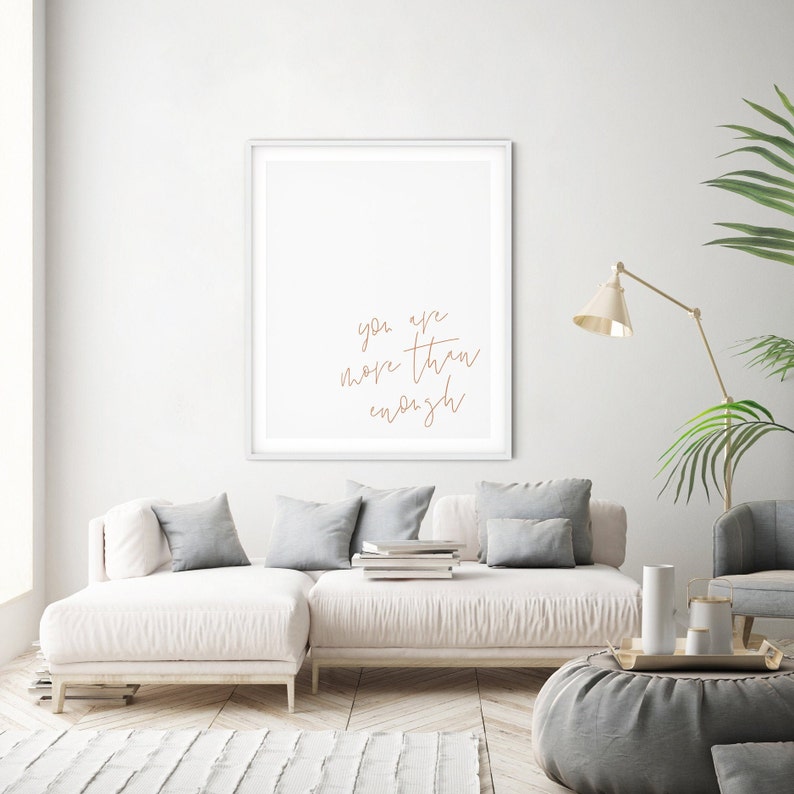 May include: A white framed print with the text "you are more than enough" in brown script on a white background. The print is hanging on a white wall above a white couch with gray pillows.