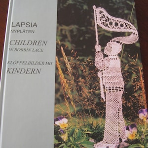 May include: Book cover with the title "Lapsia Nyplaten Children in Bobbin Lace Kloppelbilder Mit Kindern" in white text on a light gray background. The cover features a white bobbin lace design of a child holding a flag.