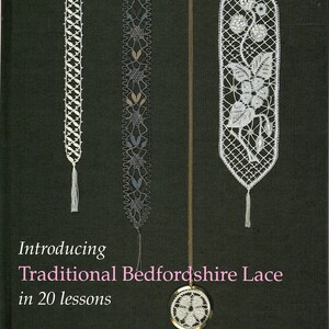May include: A book cover with the title "Introducing Traditional Bedfordshire Lace in 20 lessons" and the author's name, Barbara M Underwood. The cover features three lace bookmarks and a pendant with a lace design. The bookmarks are white and gray, with tassels.