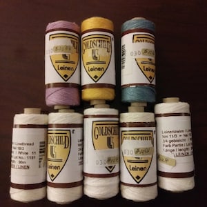 May include: Eight spools of white, yellow, blue, and pink linen thread. The spools are labeled "Goldschild Leinen" and have a number and a date on them.