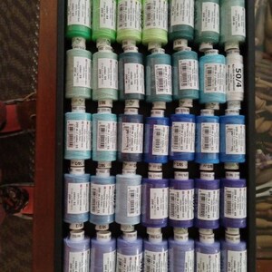 May include: A collection of 200 meter spools of thread in various shades of green, blue, and purple. The spools are arranged in a wooden box with a black frame. The spools are labeled with the color number and length of thread.