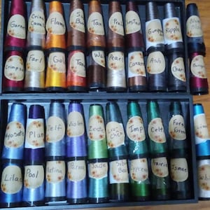 May include: A close-up view of two rows of spools of thread, each with a different color and a label with a name written on it. The spools are arranged in a wooden box with a black frame.