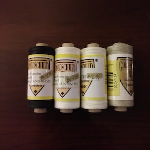 May include: Four spools of Goldschild linen thread in black, white, and off-white. Each spool has a yellow band and a label with text. The spools are arranged on a dark brown surface.