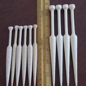 May include: A set of 11 wooden bobbins for lace making, ranging in size from small to large. The bobbins are white and have a pointed tip. They are arranged in two rows on a brown surface.