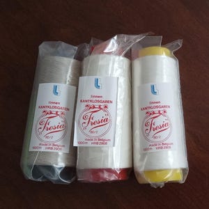 May include: Three spools of linen thread, each wrapped in clear plastic. The spools are labeled "Fresia" and "linnen KANTKLOSGAREN". The thread colors are off-white, white, and a light beige. The spools have red and yellow bases.