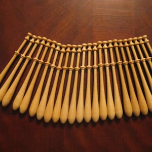 May include: A set of 25 wooden bobbins for lace making. The bobbins are arranged in a semi-circle on a brown wooden surface.