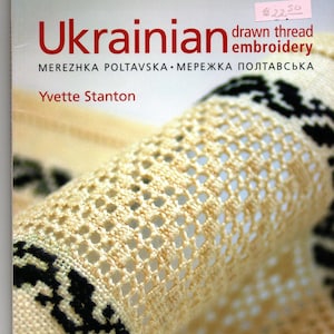 May include: A book titled Ukrainian drawn thread embroidery MEREZHKA POLTAVSKA by Yvette Stanton. The cover features a close-up of cream-colored fabric with intricate openwork drawn thread embroidery and black geometric patterns.