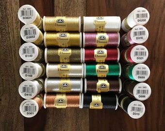 May include: A collection of DMC Diamant metallic embroidery thread spools in various colors, including gold, silver, red, and green. Each spool is labeled with "DMC Diamant" and an art number. The spools are arranged on a wooden surface.