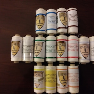 May include: A collection of 14 spools of white linen thread, each with a different color label. The labels feature the brand name "Goldschild" and the text "Leinen" which is German for linen. The spools are arranged in three rows on a wooden surface.