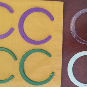 May include: An assortment of C-shaped craft stencils in various colors, including purple, green, and clear. Some stencils are on a yellow surface, while others are on a brown surface. The purple stencil has the text "PROVO LACE".