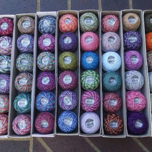 May include: A collection of 60 colorful balls of embroidery floss, arranged in 10 rows of 6 balls each. The balls are in a variety of colors, including red, blue, green, yellow, and purple. The balls are arranged in a grid pattern, and each ball is in its own individual box.
