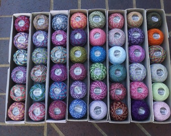 May include: A collection of 60 colorful balls of embroidery floss, arranged in 10 rows of 6 balls each. The balls are in a variety of colors, including red, blue, green, yellow, and purple. The balls are arranged in a grid pattern, and each ball is in its own individual box.