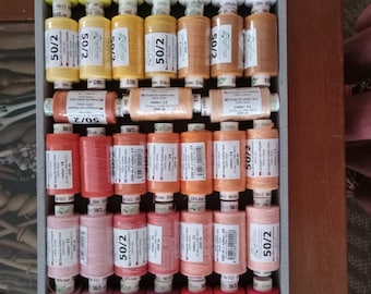 May include: A tray filled with spools of thread in various shades of yellow, orange, and pink. Each spool is labeled with "50/2" and "200 m". The threads are neatly arranged, showcasing a spectrum of colors.