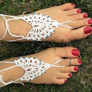 Puede incluir: Sandalias descalzas de crochet blanco con cuentas azules. Las sandalias se llevan en un par de pies con esmalte de uñas rojo.