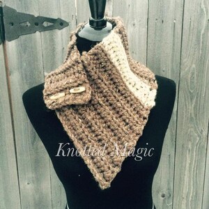May include: A brown and cream crocheted cowl with a wooden button closure. The cowl is displayed on a black mannequin against a wood background. The text "Knotted Magic" is visible below the cowl.
